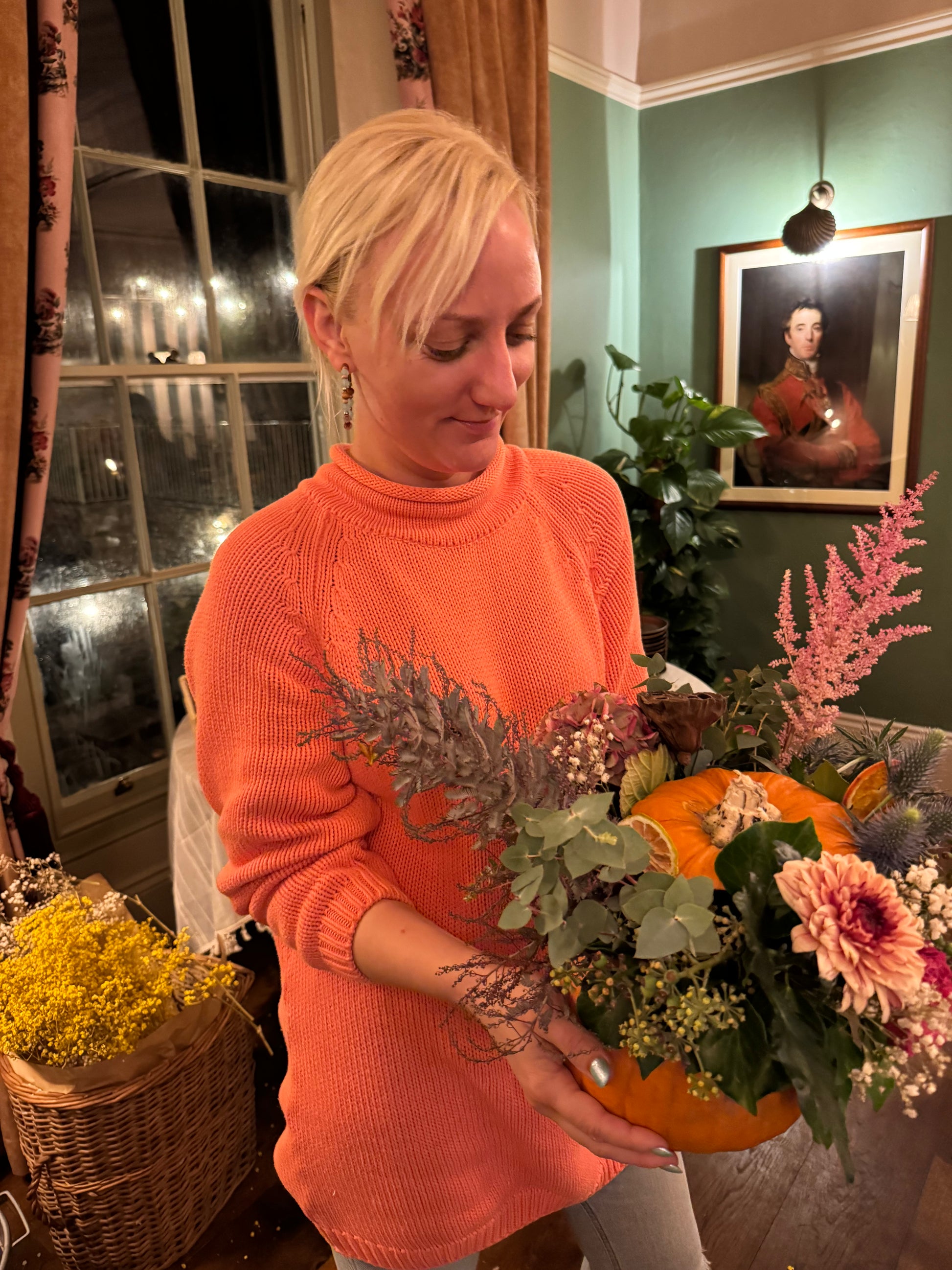 A woman with blonde hair wearing a bright orange knit turtleneck sweater holds a decorative autumn floral arrangement featuring a small orange pumpkin, pink flowers, and green foliage in an indoor setting.