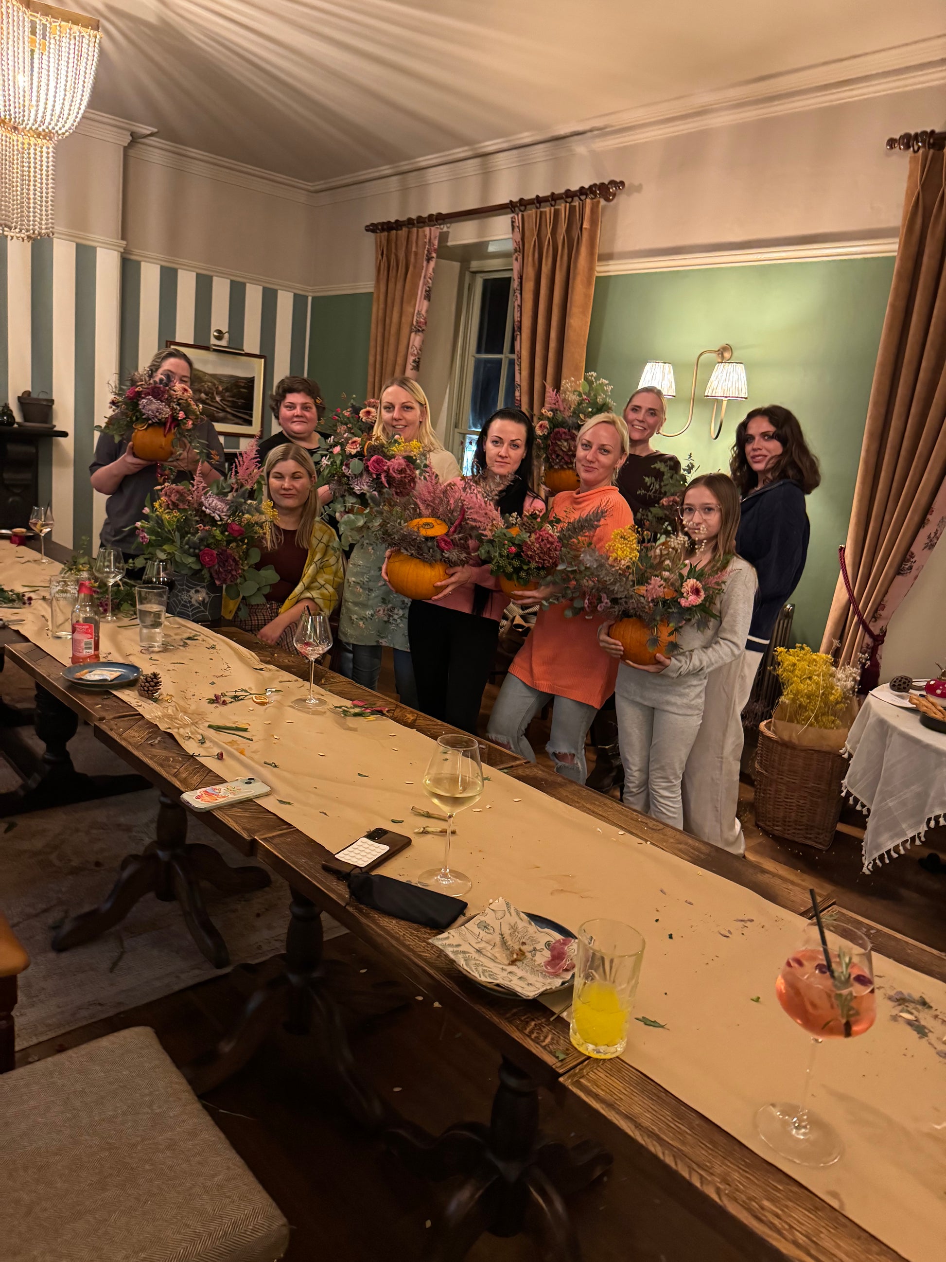 A group of women standing behind a long dining table in a decorated room, each holding a colorful floral arrangement in orange pots.