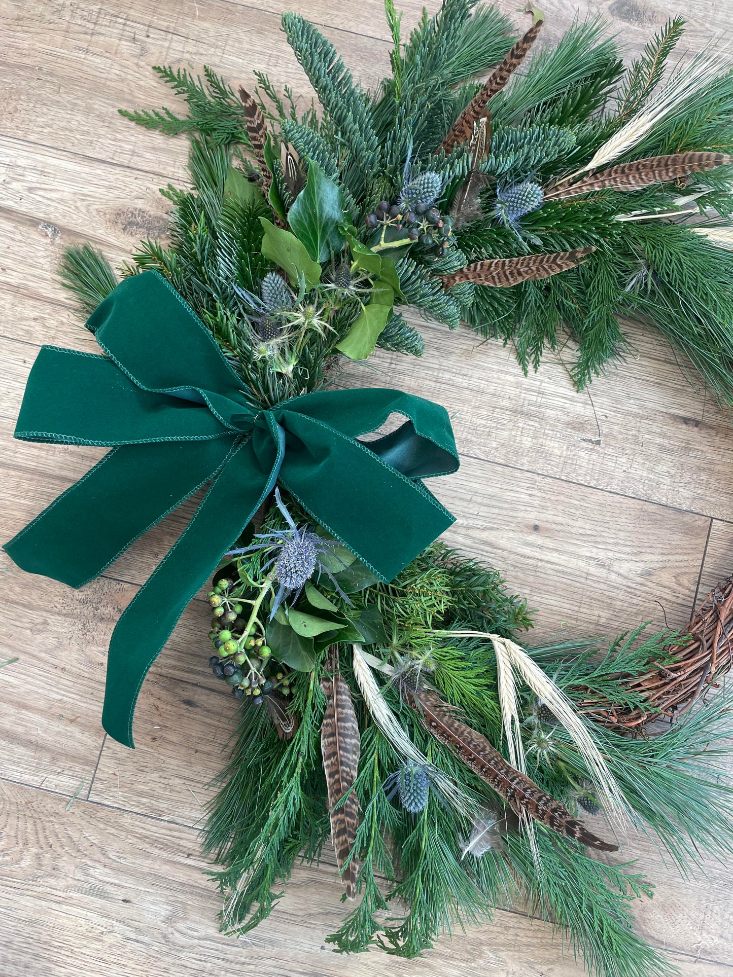 A handmade Christmas wreath made with greenery, feathers, and a green velvet ribbon bow.