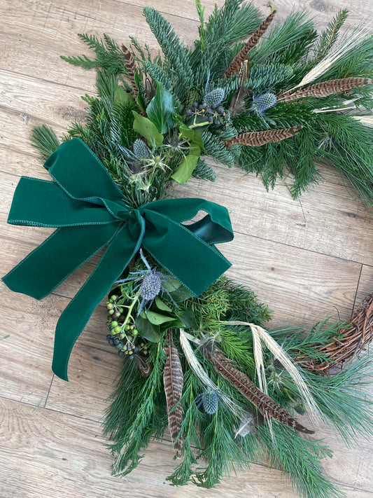 A handmade Christmas wreath made with greenery, feathers, and a green velvet ribbon bow.