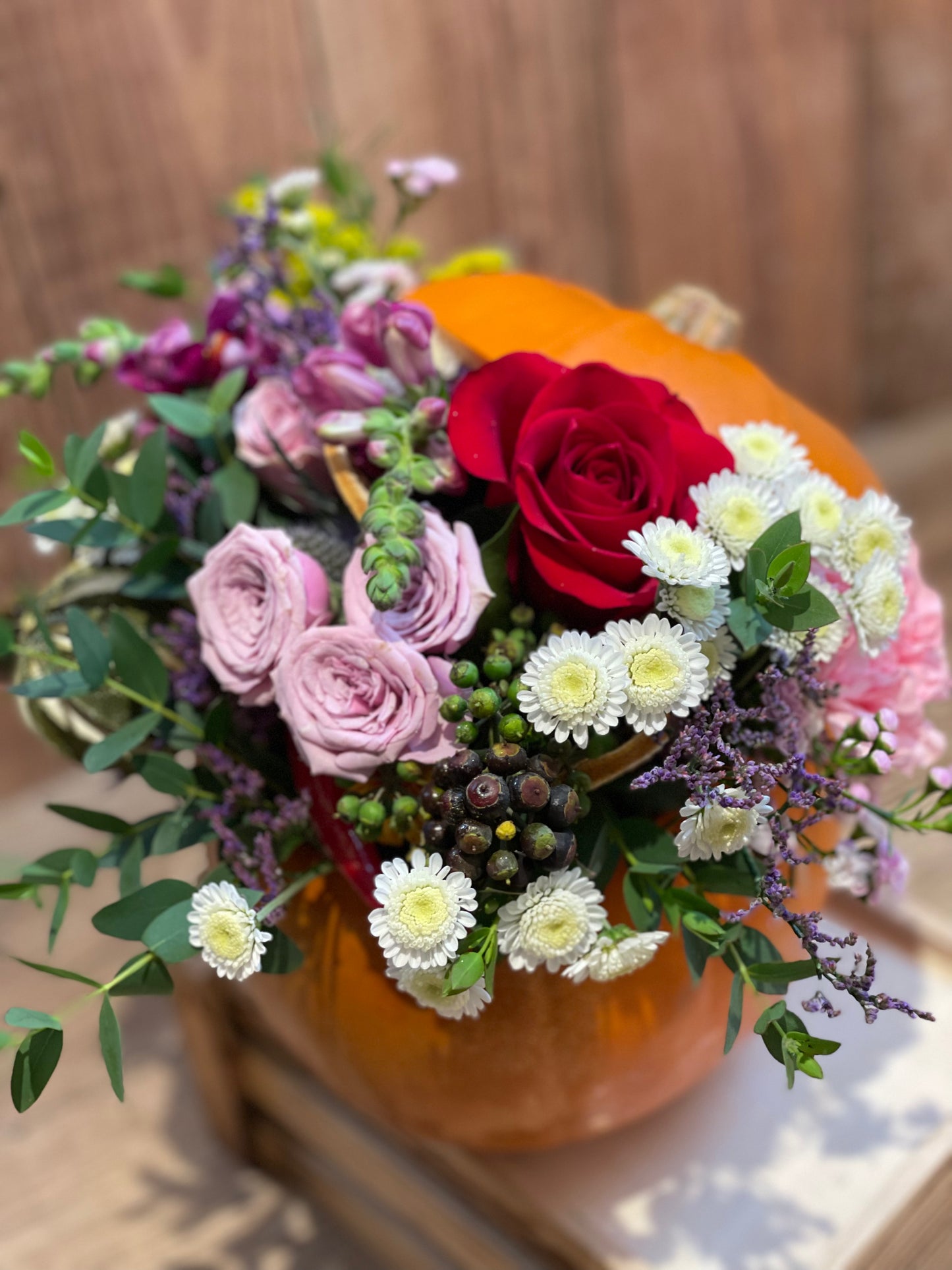 A decorative floral arrangement featuring a large red rose, light pink roses, white daisies, and green foliage, styled inside an orange pumpkin on a wooden surface.