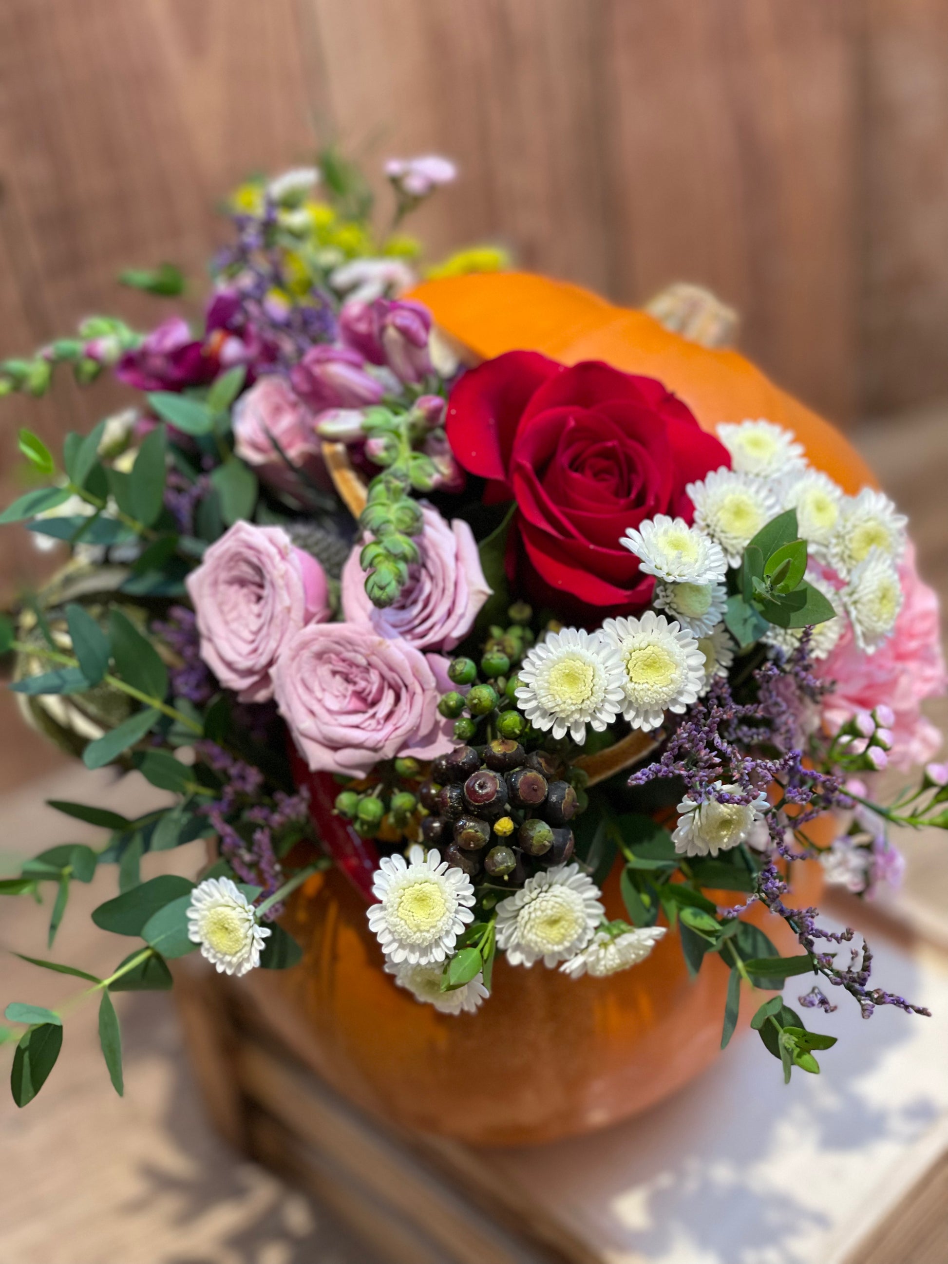 A decorative floral arrangement featuring a large red rose, light pink roses, white daisies, and green foliage, styled inside an orange pumpkin on a wooden surface.